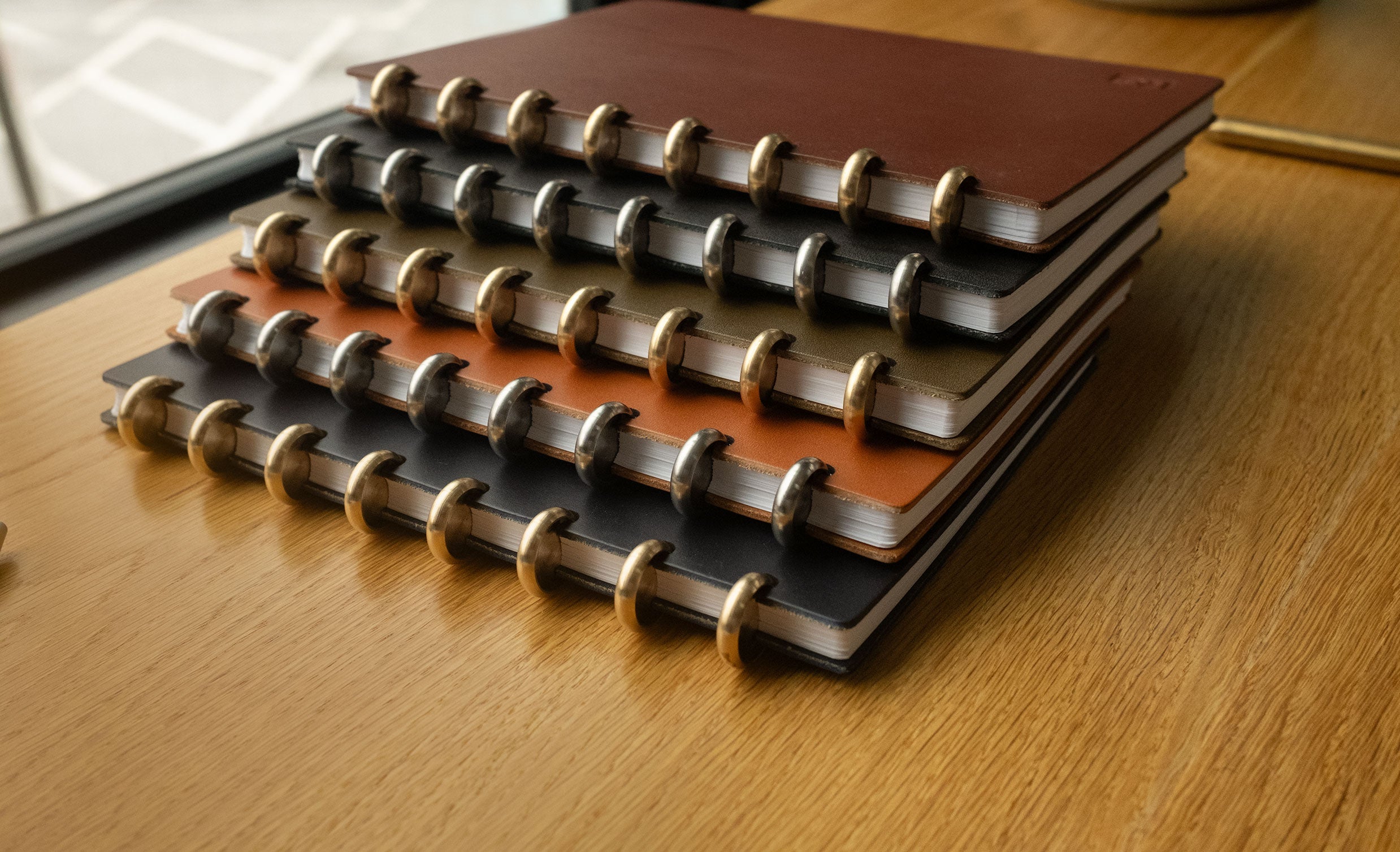 A stack of four spiral-bound notebooks with differently colored covers—brown, green, orange, and black—sits on a wooden desk near a window.