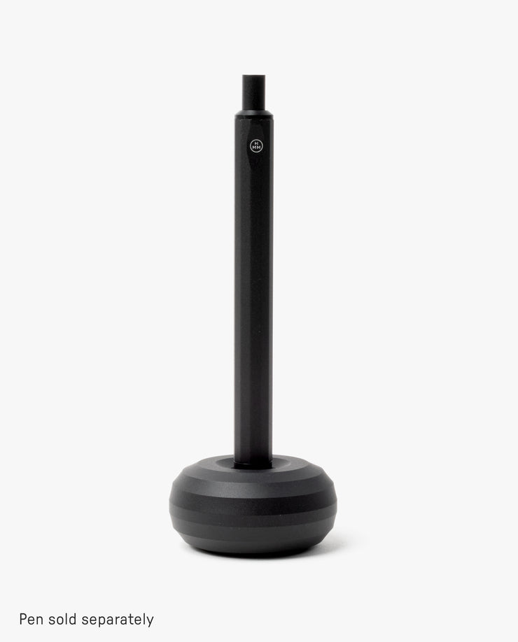 A sleek desk accessory, the HMM Paperweight Pen Stand (Black) features a round, weighted aluminum base and a vertical slot for upright pen storage. Plain white background. Text at bottom: Pen sold separately.