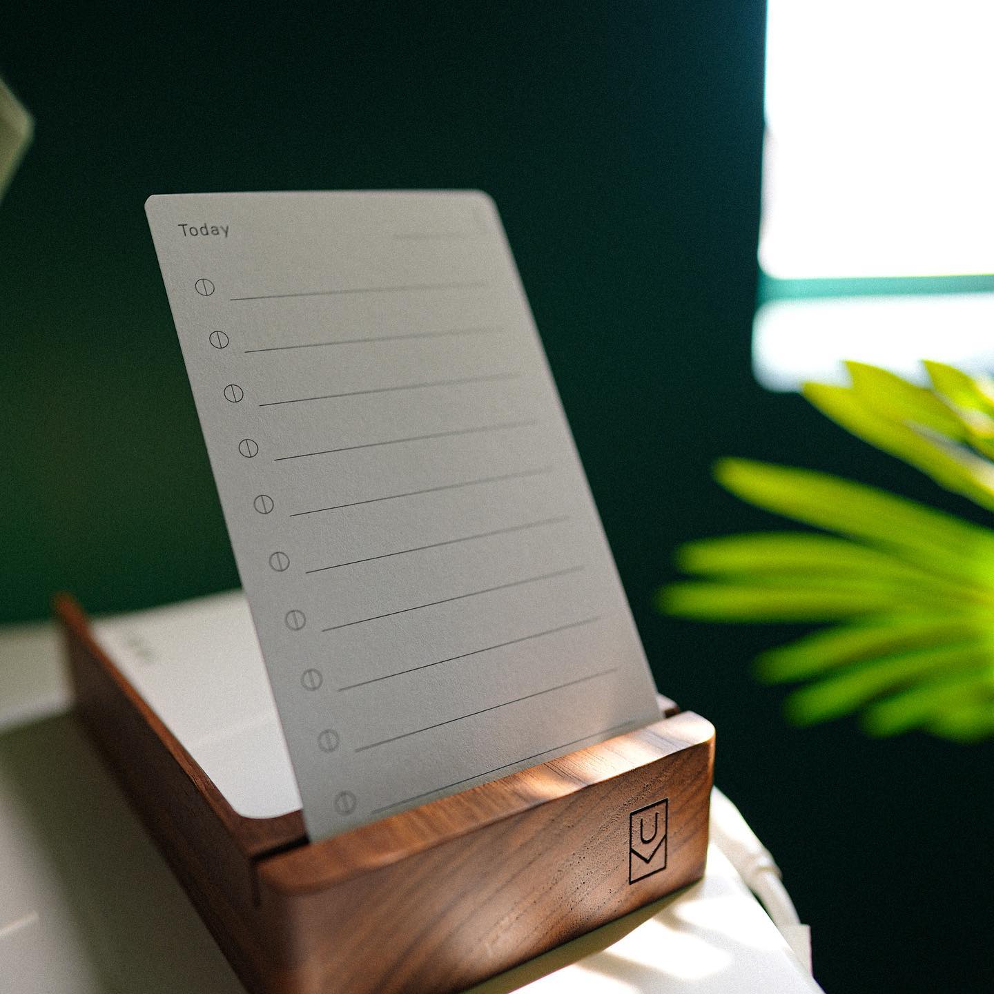 A daily planner page sits on a wooden stand, positioned on a desk near a green plant and a bright window. The planner has lines and circles for listing tasks under the heading Today.