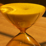Close-up of the 25 Minute Hourglass (Amber), a hand-blown glass timer with yellow sand flowing between chambers, set on a wooden surface under warm lighting.