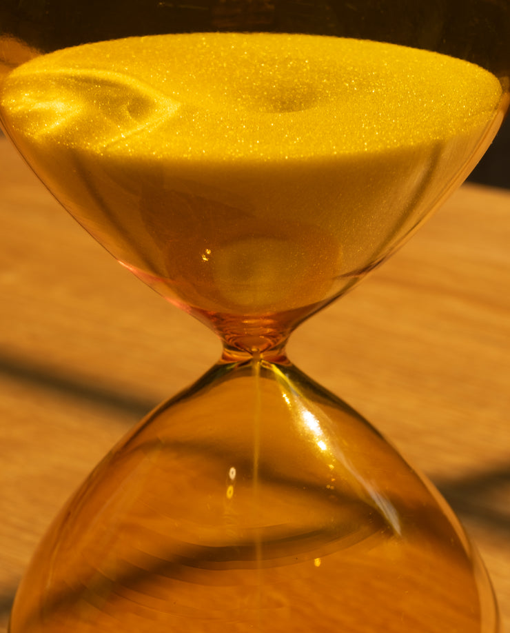 Close-up of the 25 Minute Hourglass (Amber), a hand-blown glass timer with yellow sand flowing between chambers, set on a wooden surface under warm lighting.