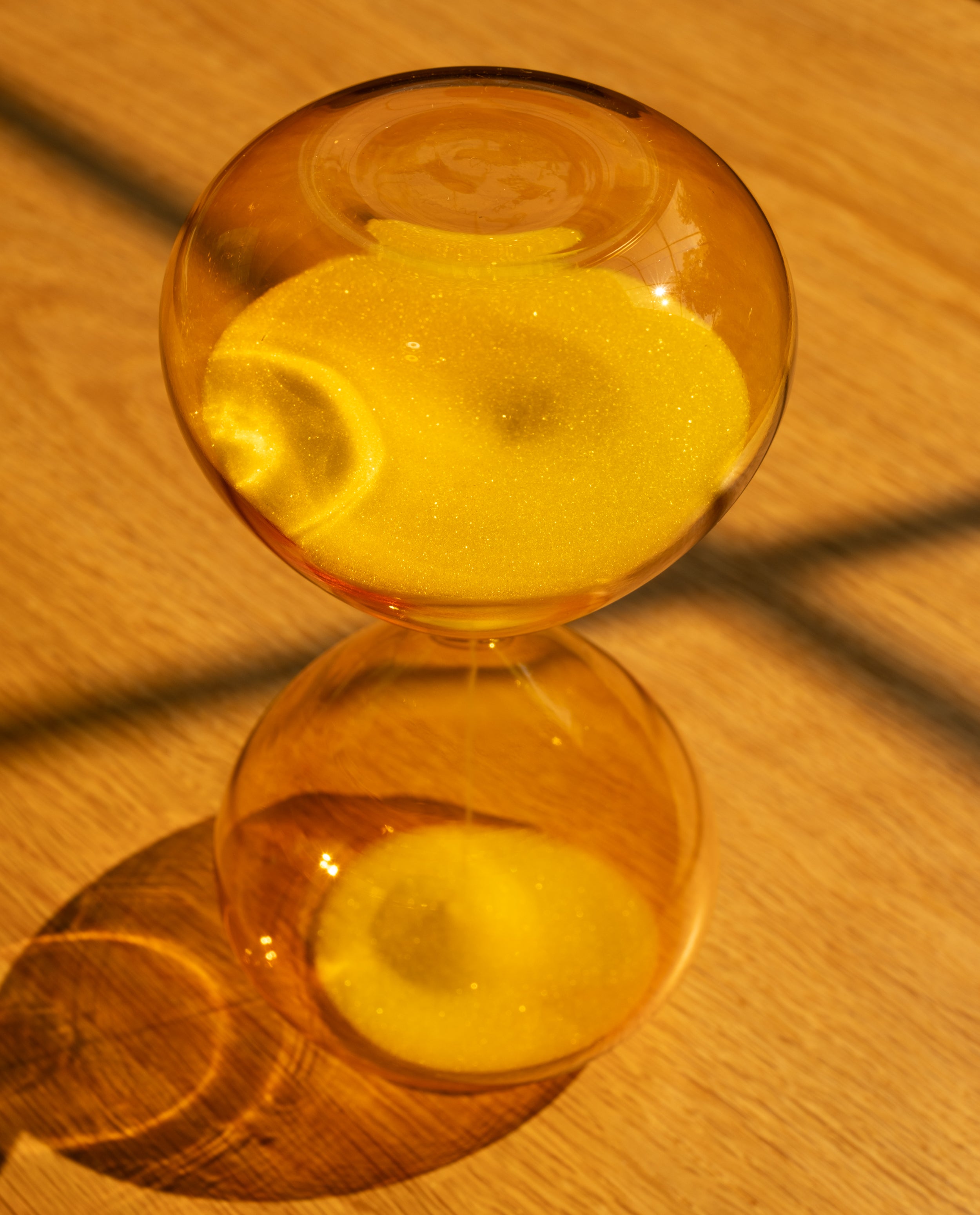 The 25 Minute Hourglass (Amber), hand-blown with yellow glass and sand, sits on a wooden surface casting a sunlit shadow. Ideal as a Pomodoro timer, its sand runs for 25 minutes to help you stay focused during work sessions.