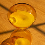 The 25 Minute Hourglass (Amber), hand-blown with yellow glass and sand, sits on a wooden surface casting a sunlit shadow. Ideal as a Pomodoro timer, its sand runs for 25 minutes to help you stay focused during work sessions.