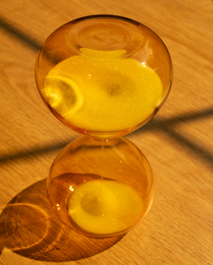 The 25 Minute Hourglass (Amber), hand-blown with yellow glass and sand, sits on a wooden surface casting a sunlit shadow. Ideal as a Pomodoro timer, its sand runs for 25 minutes to help you stay focused during work sessions.