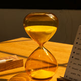 The 25 Minute Hourglass (Amber), a hand-blown glass timer with yellow sand, sits on a wooden desk in warm sunlight beside a pen and notepad.
