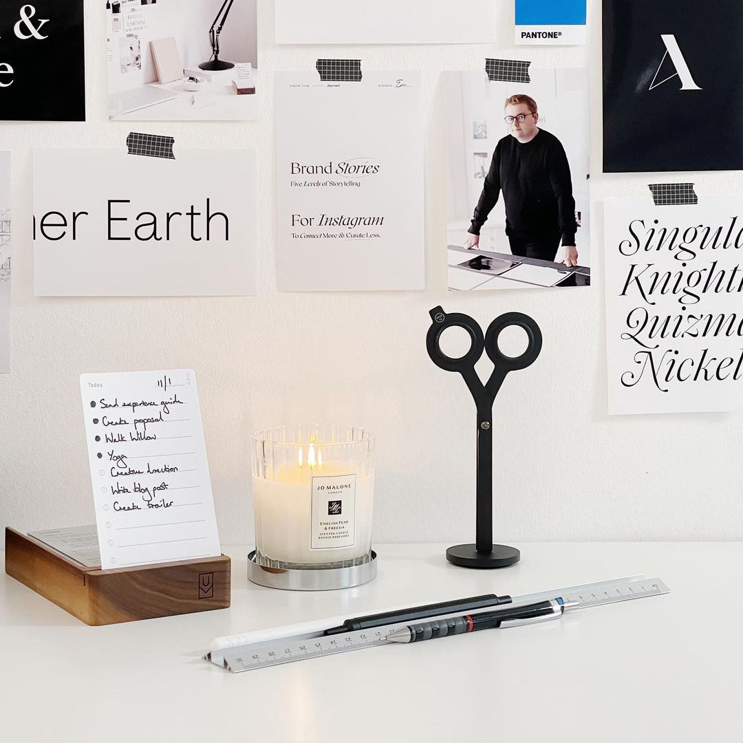 A minimalist desk setup with a candle, a ruler, a pen, scissor-shaped decor, and a notepad. The wall behind is decorated with notes, papers, and design inspiration images.