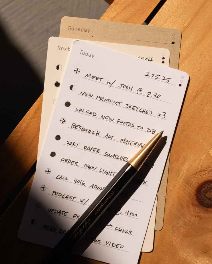 The Analog Daily Focus Kit (Walnut) displays a handwritten to-do list with tasks like meeting with Josh, product sketches, and uploading photos on a wooden surface beside a black pen and additional task cards for organized productivity.