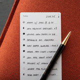 A premium Analog Trial Pack—including a handwritten to-do list on quality card stock and a black and gold pen—rests on an orange surface with tasks like meetings, research, photo uploads, and podcast recording displayed elegantly.