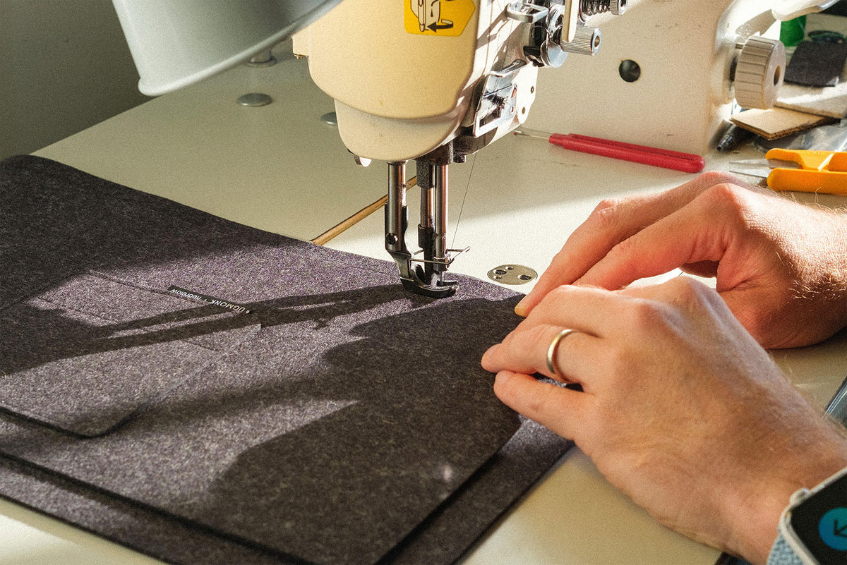 Hands guiding a piece of dark fabric through a sewing machine under bright light, with sewing tools like scissors and pencils visible on the work surface nearby.