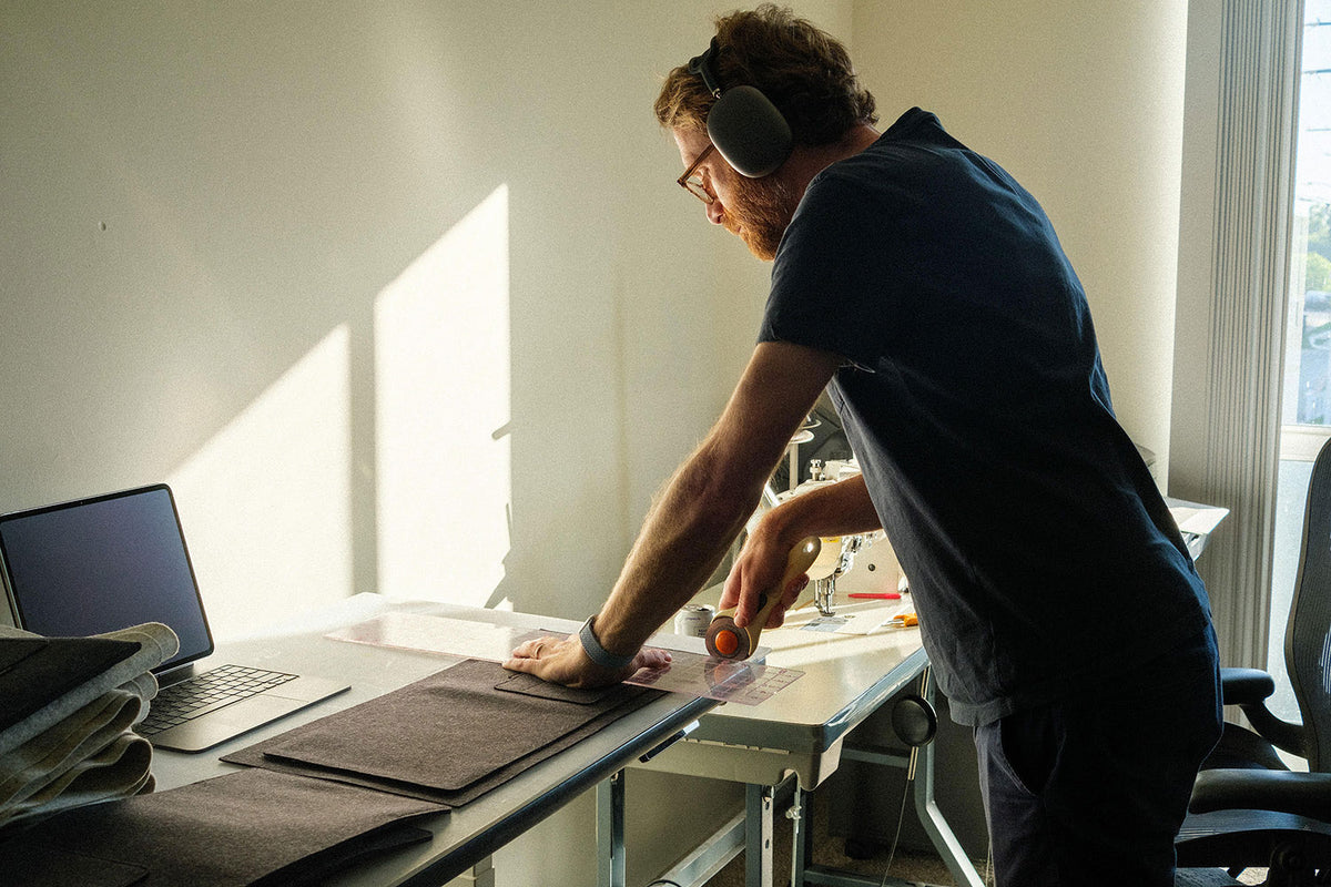 A man wearing earmuffs uses a rotary cutter to cut fabric on a worktable near a laptop in a sunlit room.