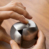 A person’s hands are holding and manipulating the Craighill Venn Puzzle (Stainless Steel), with its interlocking curved pieces forming a sphere, against a wooden surface background.