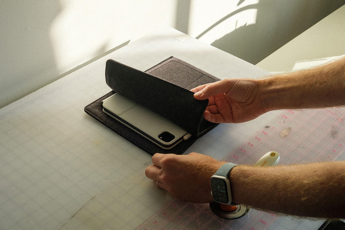 A person places a silver laptop and a smartphone into a black felt sleeve on a table, with a cutting mat and rotary cutter nearby. Sunlight casts soft shadows on the workspace.