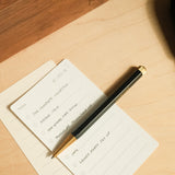 A notepad with a to-do list and The Ugmonk Pen (Leuchtturm1917 Drehgriffel - Black) placed diagonally on top, resting on a wooden desk. Tasks include The Compass Collection, Bigger Item, Copy, and Launch Party Set Up.