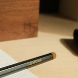 A close-up of a pencil engraved with UGMONK rests on a lined notebook atop a light wooden desk, with The Ugmonk Pen (Leuchtturm1917 Drehgriffel - Black) and a wooden box bearing a subtle logo softly blurred in the background.