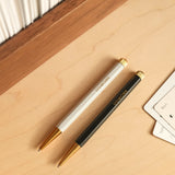 A white Ugmonk Pen and a black Ugmonk Pen (Leuchtturm1917 Drehgriffel - Black), both with gold accents and UGMONK branding, lie parallel on a light wooden desk beside paper and books.