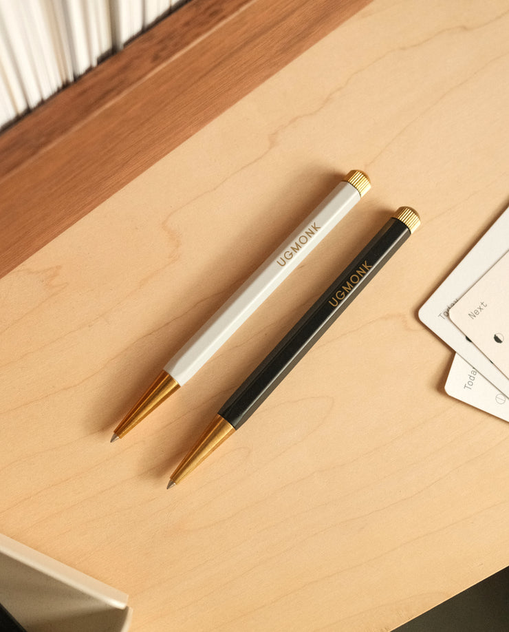 A white Ugmonk Pen and a black Ugmonk Pen (Leuchtturm1917 Drehgriffel - Black), both with gold accents and UGMONK branding, lie parallel on a light wooden desk beside paper and books.