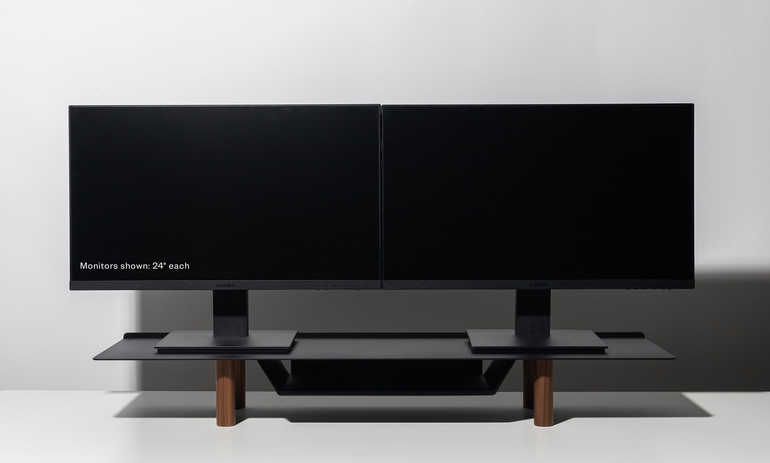 Two black 24-inch computer monitors are side by side on a Gather Large Monitor Stand (Black/Walnut) with wooden legs, set against a white background. The left monitor displays the text: “Monitors shown: 24” each.”.