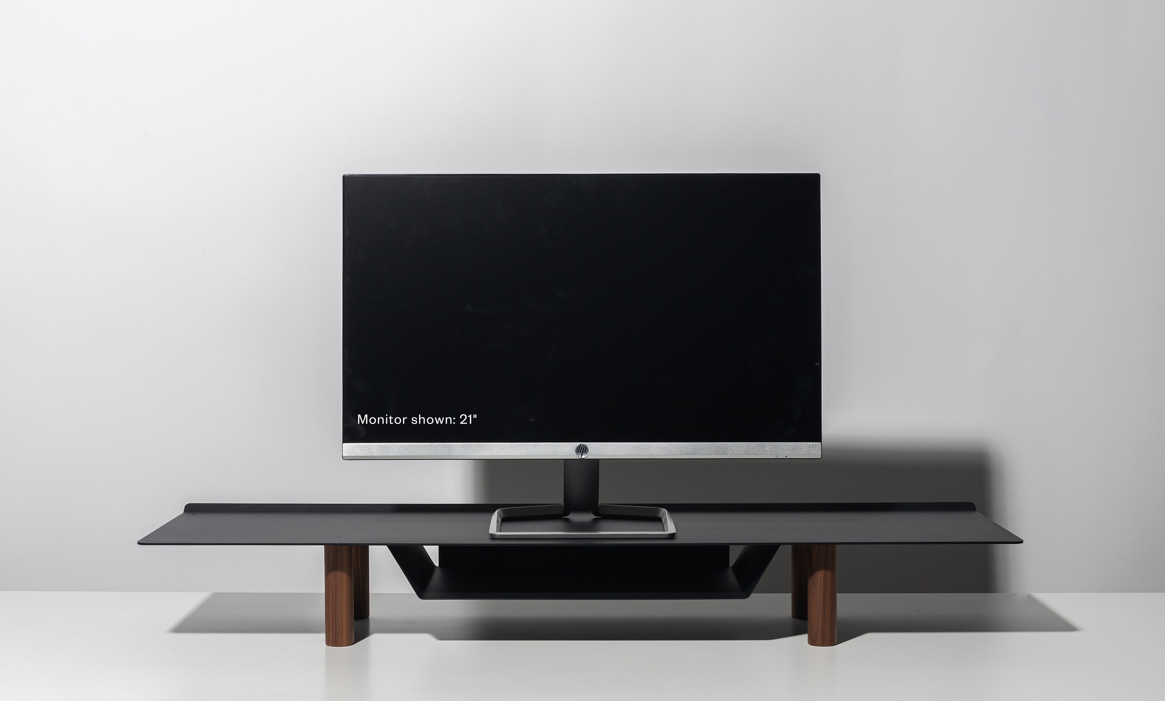 A 21-inch computer monitor sits on the Gather Large Monitor Stand (Black/Walnut) against a plain light gray background. The screen is off, drawing attention to the stand’s sleek, minimalist design and space-saving functionality.