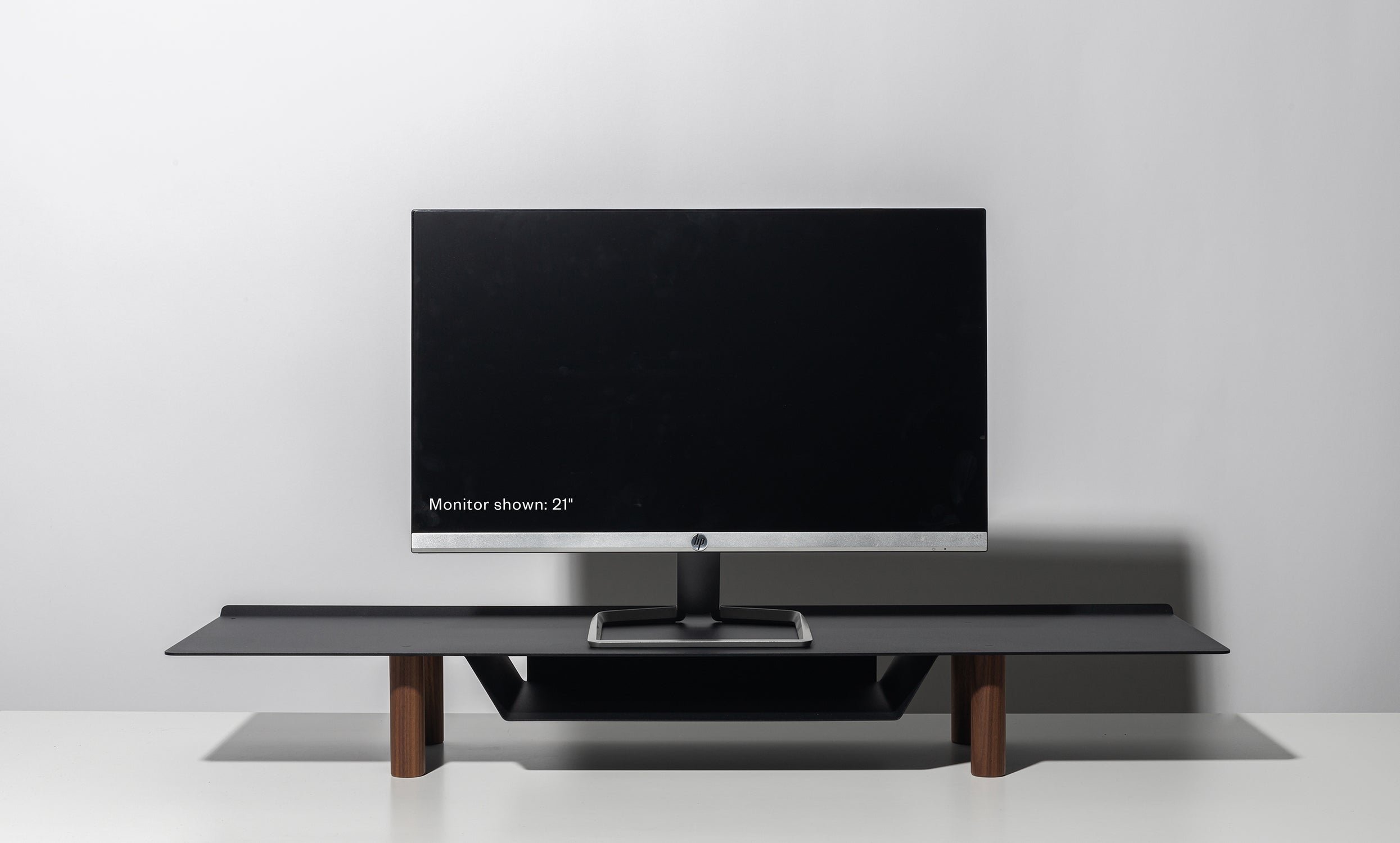 A 21-inch computer monitor sits on the Gather Large Monitor Stand (Black/Walnut) against a plain light gray background. The screen is off, drawing attention to the stand’s sleek, minimalist design and space-saving functionality.