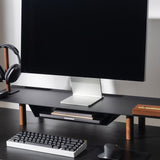 A minimalist black desk features the Gather Large Monitor Stand (Black/Walnut), with a monitor, keyboard, mouse, headphones on a holder, pen, and a compact desk organizer for stationery, all neatly arranged on a clean workspace.
