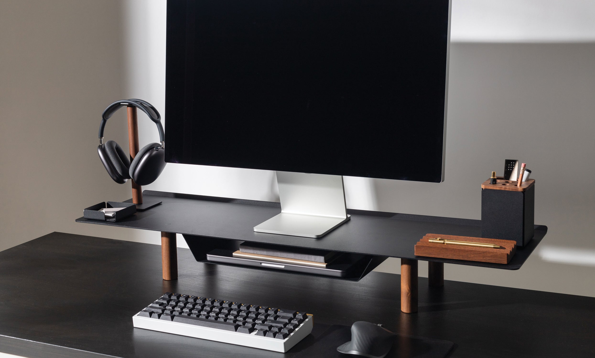 A minimalist black desk features the Gather Large Monitor Stand (Black/Walnut), with a monitor, keyboard, mouse, headphones on a holder, pen, and a compact desk organizer for stationery, all neatly arranged on a clean workspace.