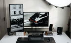A modern, minimalist desk setup features a vertical monitor on the Gather Large Monitor Stand (Black/Walnut) displaying a black-and-white street photo, a wide monitor with a vintage car image, plus keyboard, mouse, speakers, and neatly arranged tech accessories.