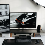 A modern, minimalist desk setup features a vertical monitor on the Gather Large Monitor Stand (Black/Walnut) displaying a black-and-white street photo, a wide monitor with a vintage car image, plus keyboard, mouse, speakers, and neatly arranged tech accessories.