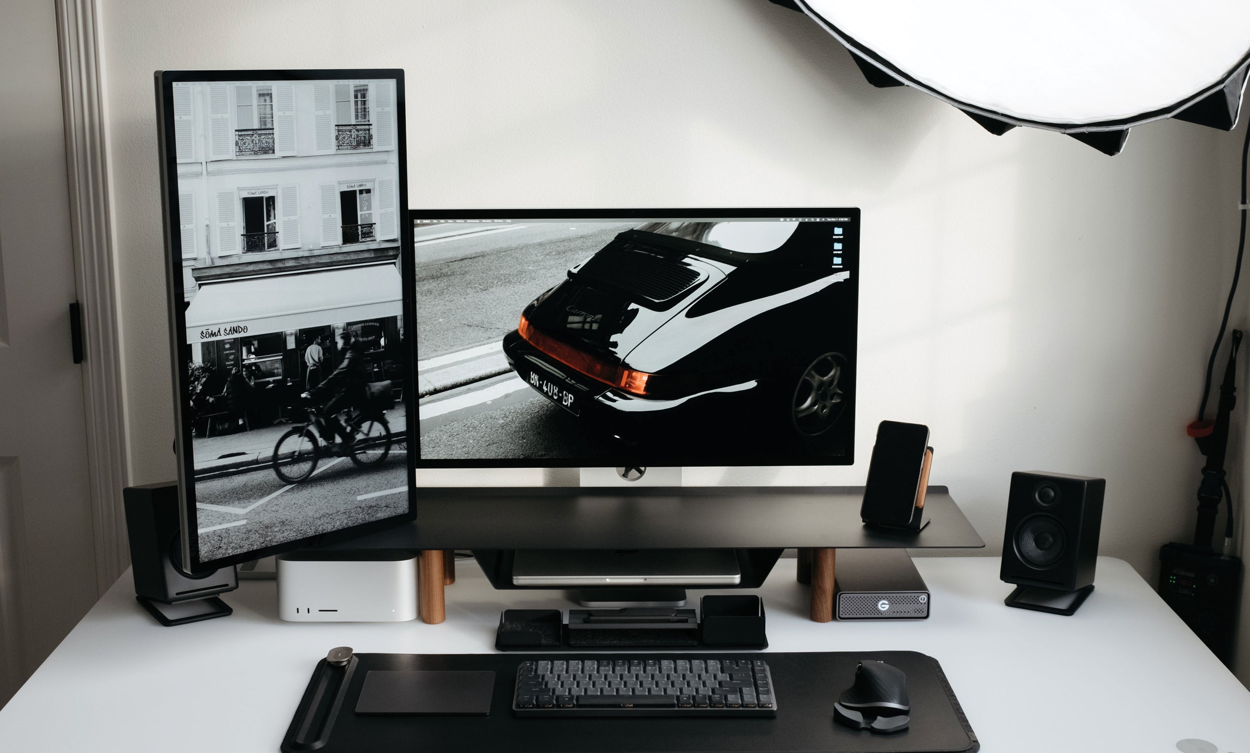 A modern, minimalist desk setup features a vertical monitor on the Gather Large Monitor Stand (Black/Walnut) displaying a black-and-white street photo, a wide monitor with a vintage car image, plus keyboard, mouse, speakers, and neatly arranged tech accessories.