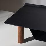 A close-up of the Gather Small Monitor Stand (Black/Walnut), featuring a sleek black powder-coated steel top and a single cylindrical walnut leg, set against a neutral background with soft lighting and shadows.