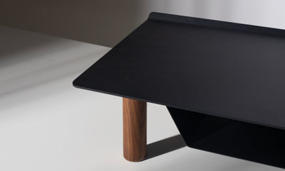 A close-up of the Gather Small Monitor Stand (Black/Walnut), featuring a sleek black powder-coated steel top and a single cylindrical walnut leg, set against a neutral background with soft lighting and shadows.