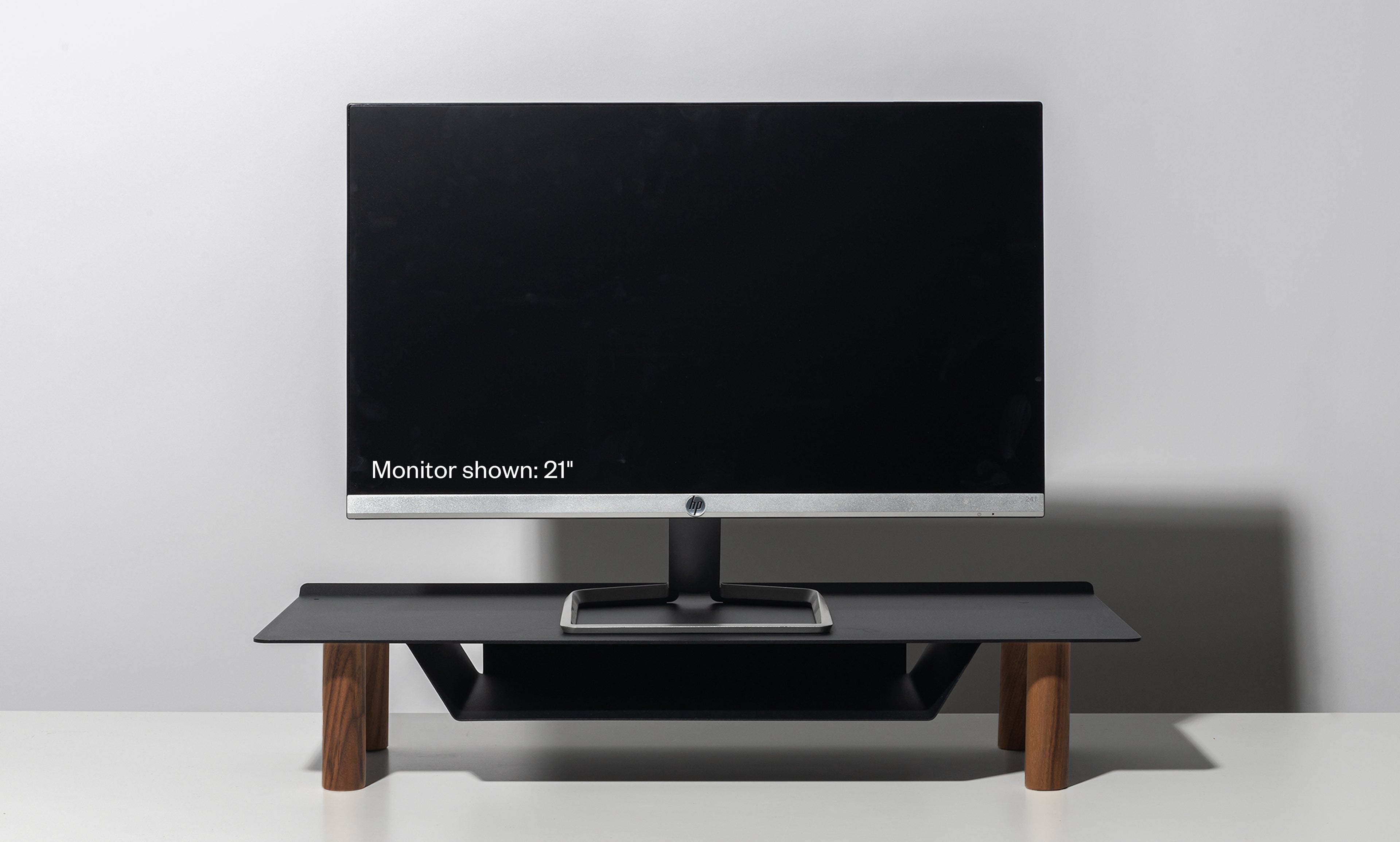 A 21-inch computer monitor rests on the Gather Small Monitor Stand (Black/Walnut) with wooden legs, set against a plain light gray background. The screen is off, displaying the text: “Monitor shown: 21”.”.