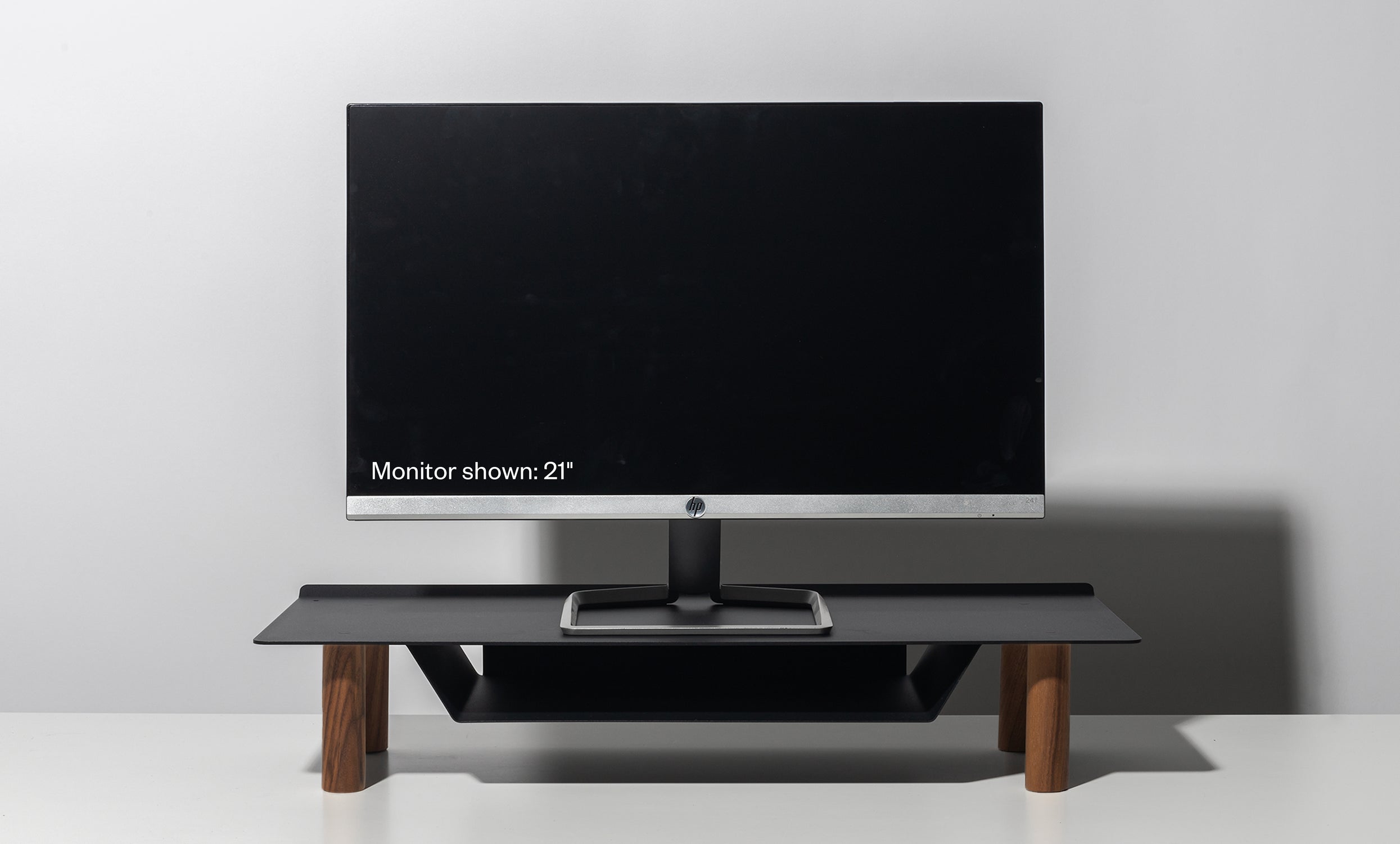 A 21-inch computer monitor rests on the Gather Small Monitor Stand (Black/Walnut) with wooden legs, set against a plain light gray background. The screen is off, displaying the text: “Monitor shown: 21”.”.