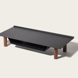 The Gather Small Monitor Stand (Black/Walnut) features a black rectangular top, four cylindrical walnut legs, and a built-in black powder-coated steel storage shelf, displayed against a plain light background.