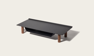 The Gather Small Monitor Stand (Black/Walnut) features a black rectangular top, four cylindrical walnut legs, and a built-in black powder-coated steel storage shelf, displayed against a plain light background.