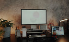 A modern workspace features a computer monitor elevated on the Gather Small Monitor Stand (White/Walnut), alongside a Mac Mini, keyboard, mouse, tablet, smartphone, potted plant, pens, and glasses arranged neatly on a dark desk against a textured wall. The monitor displays overlapping circles.