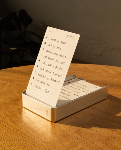 The Analog Daily Focus Kit + 6 Refills (Silver Aluminum) holds a handwritten to-do list in its minimalist card holder on a wooden table, bathed in warm sunlight, with additional task cards neatly stacked inside.