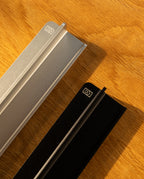 Two metal objects, one silver and one black with geometric logos on their ends, rest on wood. Warm light casts shadows, evoking the sleek design of the Analog Weekly Planning Kit (Silver Aluminum).