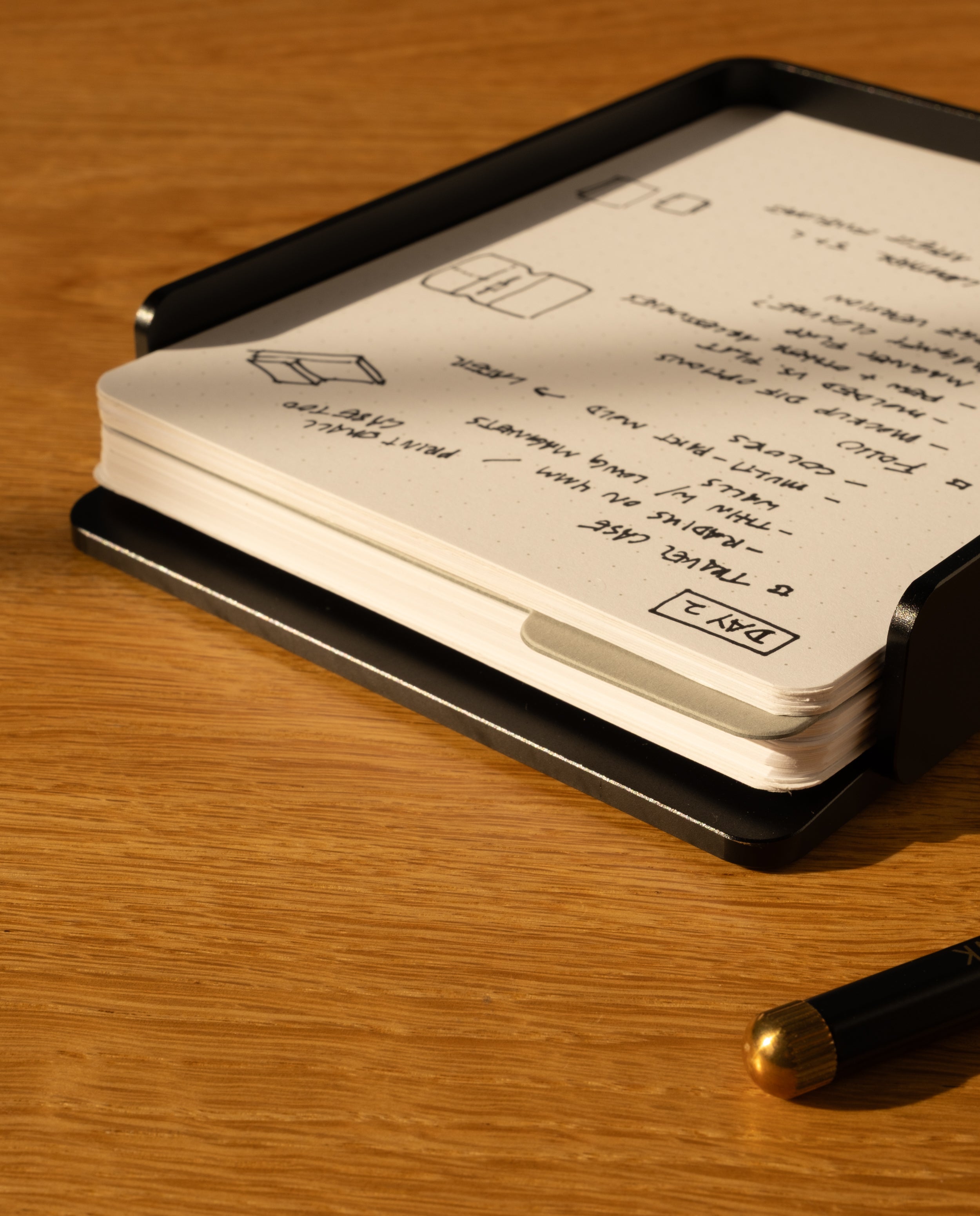 The Analog Capture Kit - Large (Black Aluminum) includes large dot grid cards in a sleek black holder, with a black pen featuring a gold tip, all resting on a wooden surface—an ideal setup for your notes.