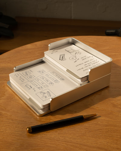 The Complete Analog Collection (Silver Aluminum) neatly holds stacked note cards with sketches and notes on a wooden desk beside a black and gold pen, while warm light casts gentle shadows across the scene.