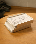 A silver aluminum Analog Capture Kit - 2 Large sits slightly open on a wooden table, with illustrated instructions on the lid. Dot grid cards and an aluminum tray are visible inside, and a leafy plant appears in the background.
