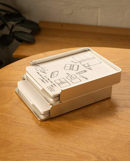 A silver aluminum Analog Capture Kit - 2 Large sits slightly open on a wooden table, with illustrated instructions on the lid. Dot grid cards and an aluminum tray are visible inside, and a leafy plant appears in the background.