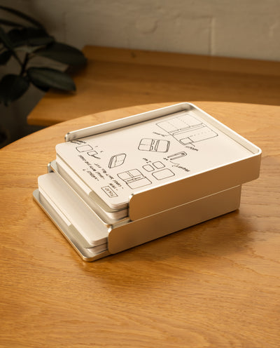 A silver aluminum Analog Capture Kit - 2 Large sits slightly open on a wooden table, with illustrated instructions on the lid. Dot grid cards and an aluminum tray are visible inside, and a leafy plant appears in the background.