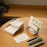 A wooden table displays The Complete Analog Collection (Silver Aluminum), organizing notecards, pens, and markers. Some cards show handwritten notes and sketches, with a plant partially visible in the background.