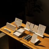 The Complete Analog Collection (Silver Aluminum) is displayed on a wooden table with neatly arranged pens, notepads, and a card organizer holding sketched white cards, all bathed in warm sunlight with a potted plant and dark furniture in the background.