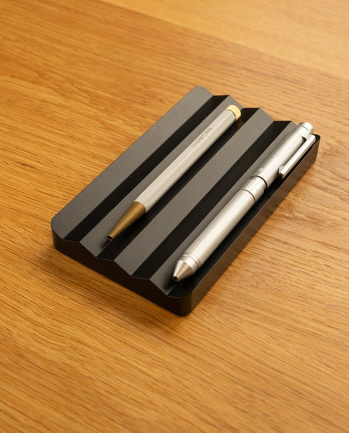 The Complete Analog Collection (Black Aluminum) features two metallic pens resting parallel on a black grooved holder atop a light wooden surface—a sleek, modern desktop organizer for your workspace.
