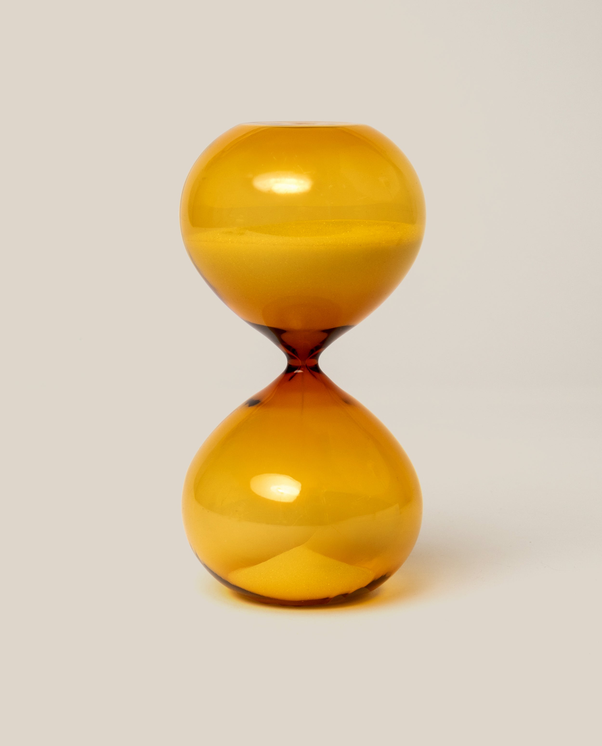 The 15 Minute Hourglass (Amber) features a transparent orange design filled with yellow sand and sits elegantly on a plain light background.