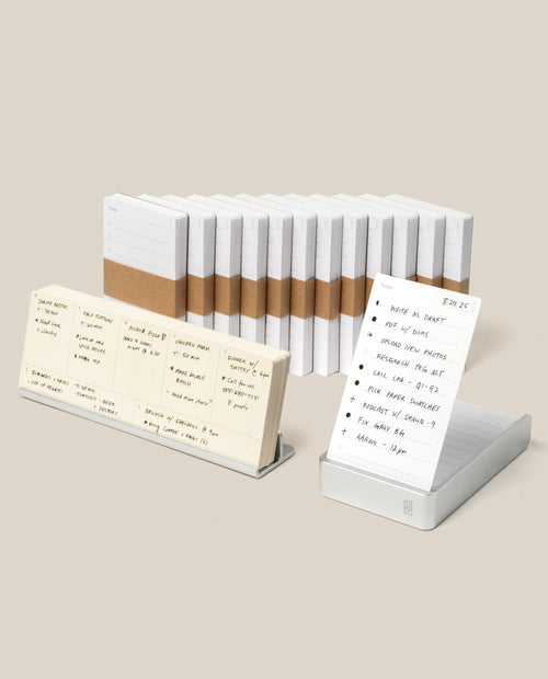 The Analog Annual Kit (Silver Aluminum) features white notepads with brown paper bands arranged in a row. Two notepads displayed on stands, like daily card holders, showcase handwritten notes against a plain beige background.