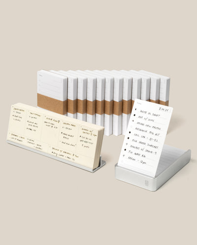The Analog Annual Kit (Silver Aluminum) features white notepads with brown paper bands arranged in a row. Two notepads displayed on stands, like daily card holders, showcase handwritten notes against a plain beige background.