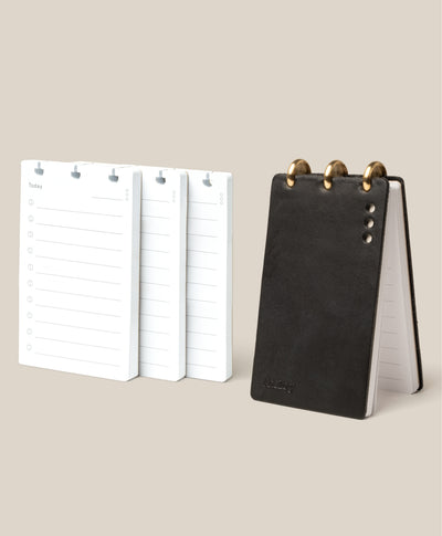 The Leather Discbound Analog + 3 Refills (Black), a sleek full-grain leather binder, stands upright beside three stacks of lined and checkboxed planner refills—ideal for organizing your Analog Task Cards.