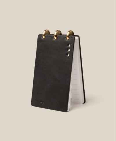The Leather Discbound Analog (Black) is a small black flip notepad with three large gold rings at the top and three small embossed dots on its full-grain leather cover, shown standing upright on a beige background.
